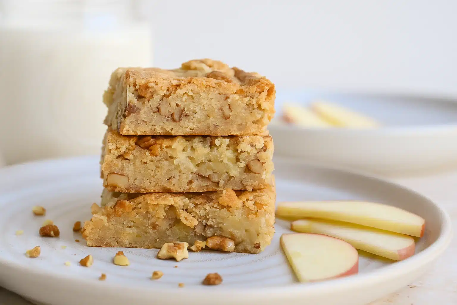 A close-up of stacked apple brownies with chopped walnuts on a ceramic plate, surrounded by apple slices and soft natural light.