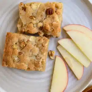 A close-up of stacked apple brownies with chopped walnuts on a ceramic plate, surrounded by apple slices and soft natural light.