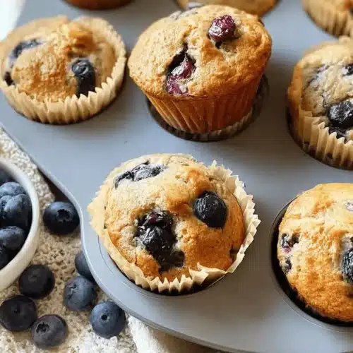 Freshly baked Blueberry Cottage Cheese Muffins in a tin, surrounded by raw blueberries, on a textured placemat.