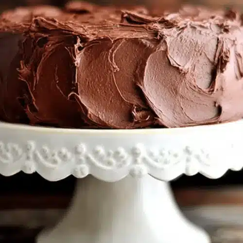 A decadent, richly frosted round chocolate cake on an elegant scalloped cake stand, perfect for a beloved chocolate cake recipe.