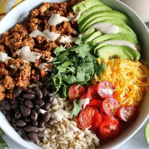 A colorful Easy Taco Rice Bowl – a quick and healthy dinner idea – filled with seasoned ground meat, avocado, cheese, tomatoes, black beans, and fresh cilantro, on a light background with lime and avocado.