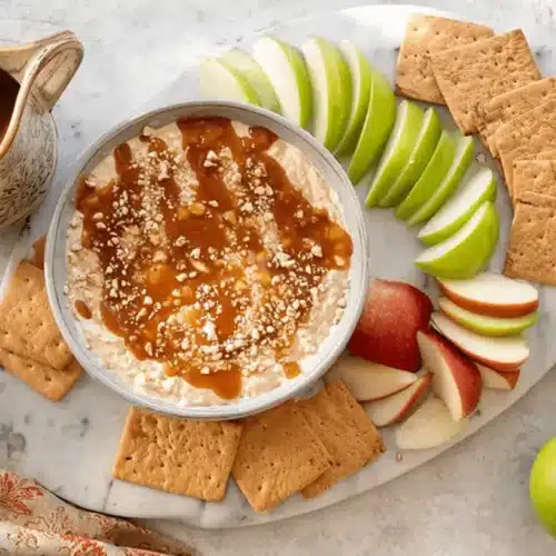 Overhead view of creamy Cream Cheese Caramel Apple Dip with caramel drizzle, nuts, sliced red and green apples, and graham crackers on a marble board.
