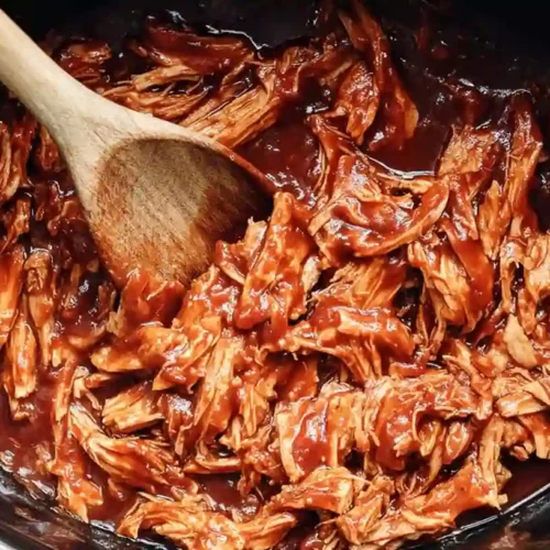 Shredded barbecue chicken cooked in a slow cooker, coated in rich BBQ sauce, and mixed with a wooden spoon—perfect for sandwiches, bowls, or tacos