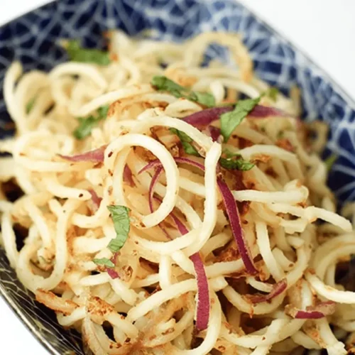 Close-up of a vibrant dish of Easy Potato Noodles with red onion, green herbs, and crispy bits in a blue and white patterned bowl.