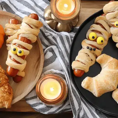 A festive Halloween party food spread on a wooden table, featuring mummy hot dogs, ghost and bat-shaped pastries, and dipping sauces, illuminated by candlelight.