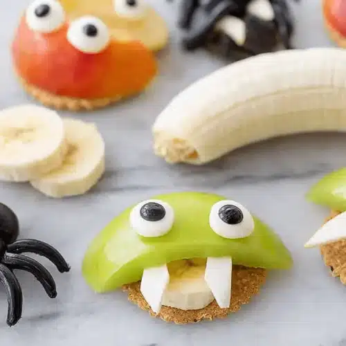 Whimsical apple monsters with candy eyes and fangs, arranged on crackers, showcasing creative and healthy Halloween snack ideas.