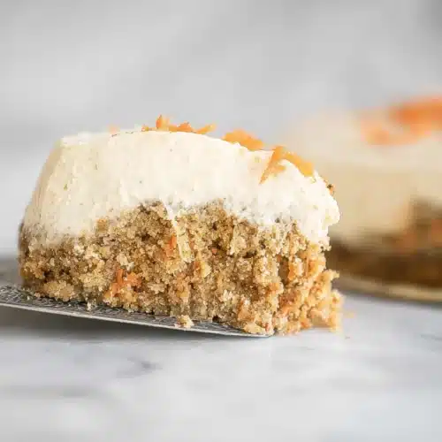 A slice of carrot cake cheesecake with a moist carrot-spice cake base, silky cheesecake layer, and a fluffy cream topping, garnished with shredded carrots.