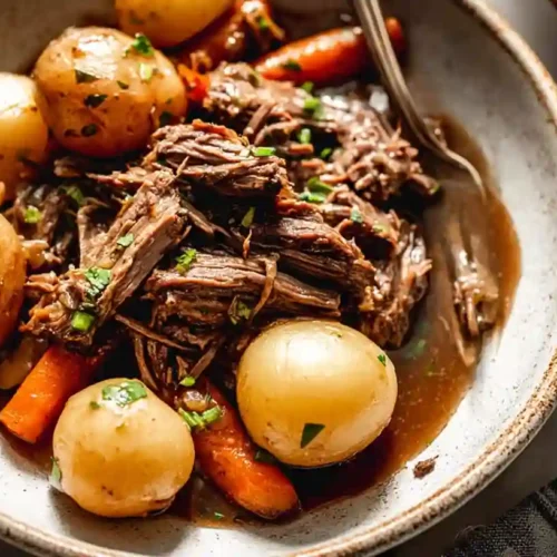 Bowl of tender shredded pot roast with baby potatoes and carrots in rich brown gravy, garnished with parsley.