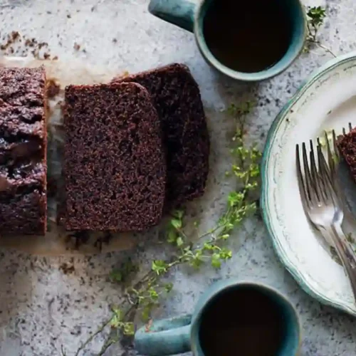 Moist chocolate loaf cake sliced on a board, with one slice on a plate and two coffee cups nearby.