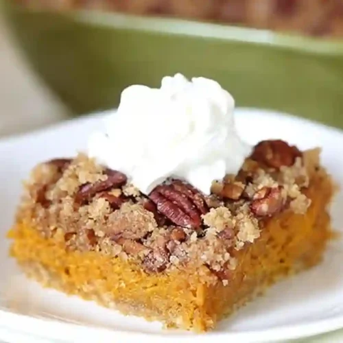 Square of pumpkin dump cake topped with buttery pecan streusel and a swirl of whipped cream on a white plate.