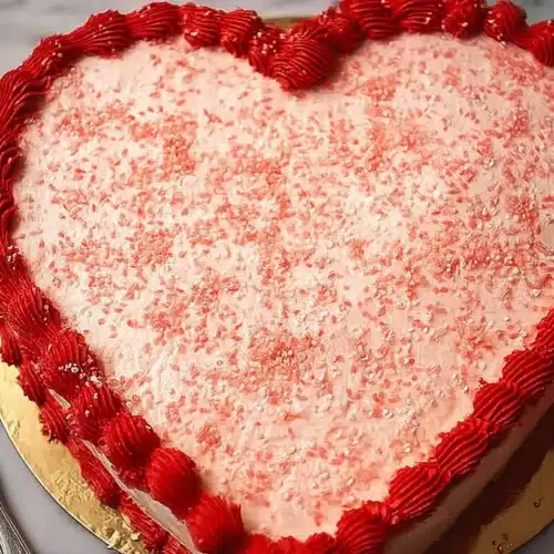 Heart‑shaped Valentine’s cake with pink frosting, red buttercream border, and pink sugar sprinkles on a gold cake board.
