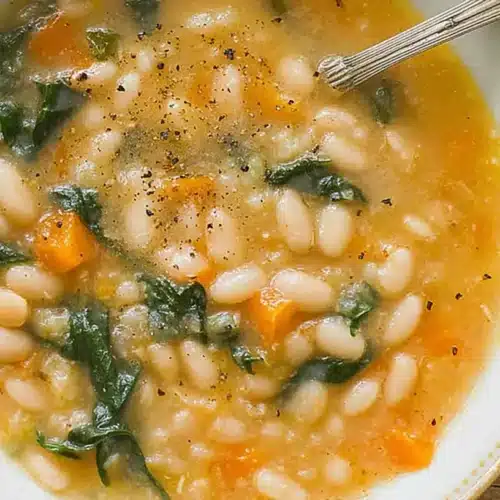Overhead view of a comforting bowl of the Best White Bean Soup, featuring creamy white beans, vibrant green leafy vegetables, and carrots on a rustic wooden table.