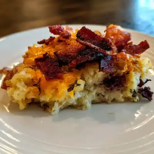 A close-up of a hearty slice of Cracked Out Tater Tot Breakfast Casserole, topped with crispy bacon and gooey melted cheddar cheese, served on a white plate.