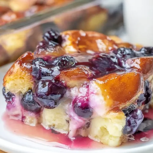 Close-up of a glazed slice of Overnight Blueberry French Toast Casserole, rich with burst blueberries, served on a white plate.