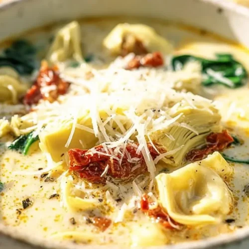 A close-up of a rustic ceramic bowl filled with Creamy Lemony Tuscan Artichoke Soup, featuring pasta, artichoke hearts, sun-dried tomatoes, spinach, and a generous topping of grated Parmesan cheese.