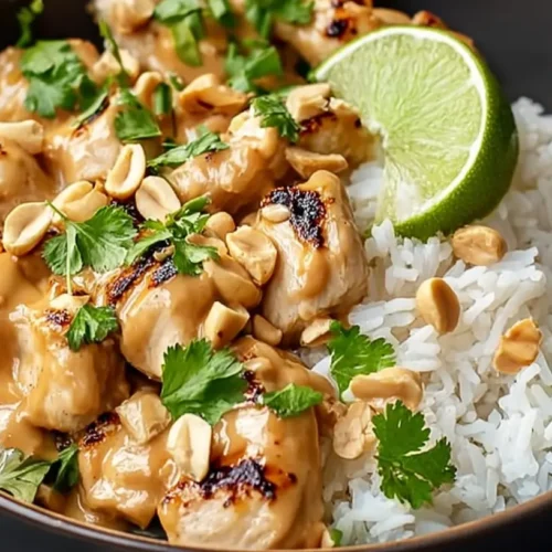 Close-up of a serving of Irresistible Thai Peanut Chicken in a dark bowl, garnished with peanuts, cilantro, and a lime wedge.